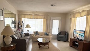 Una sala de estar con sofá, sillón, mesa de centro, televisor, lámpara y cortinas.