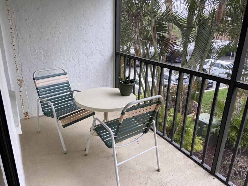 Een balkon met twee metalen stoelen en een kleine ronde tafel, een bloempot en uitzicht op palmbomen en geparkeerde auto's.