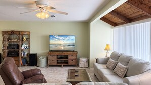 A living room with a ceiling fan, a television, a wicker entertainment center, a leather sofa, and a glass coffee table. - Bayside 2BR Condo at Tamarind - Private Sun Deck (Englewood)