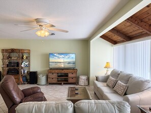 Ein Wohnzimmer mit Deckenventilator, Fernseher, einem Korbmöbel-Entertainment-Center, einer Ledersofa und einem Glastisch.
