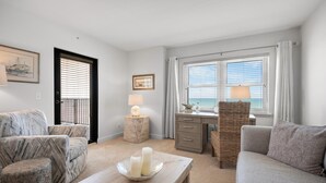A living room with a sofa, a coffee table with candles, a desk with a lamp, and a view of the sea through the window.