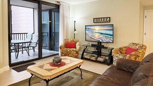 Sala de estar con televisor, dos sillones estampados, un sofá, un ventilador de techo y un ventanal corredizo que da a un área exterior con una mesa y sillas.