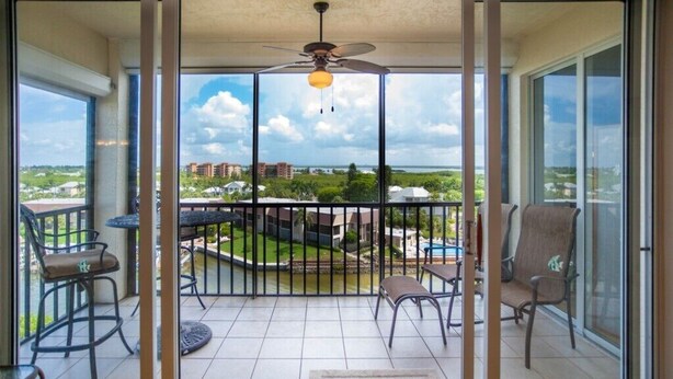 A balcony with a view of a swimming pool and buildings, furnished with chairs and a table. - 2BR – Penthouse, Pool, Beach & Water Views (Fort Myers Beach)