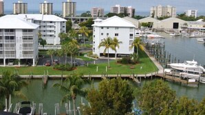 A marina with several boats docked, surrounded by modern buildings with large windows and a grassy area with palm trees. - 2BR – Penthouse, Pool, Beach & Water Views (Fort Myers Beach)
