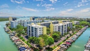 A waterfront residential complex with multiple high-rise buildings, a marina with boats, and a clear view of the city skyline in the background. - 2BR – Penthouse, Pool, Beach & Water Views (Fort Myers Beach)