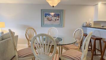 A dining area with a round glass table surrounded by six striped chairs, a chandelier, and a framed painting on the wall.