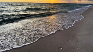 Une plage où les vagues viennent doucement lécher le rivage, un ciel dégagé et le soleil qui se couche à l’horizon.