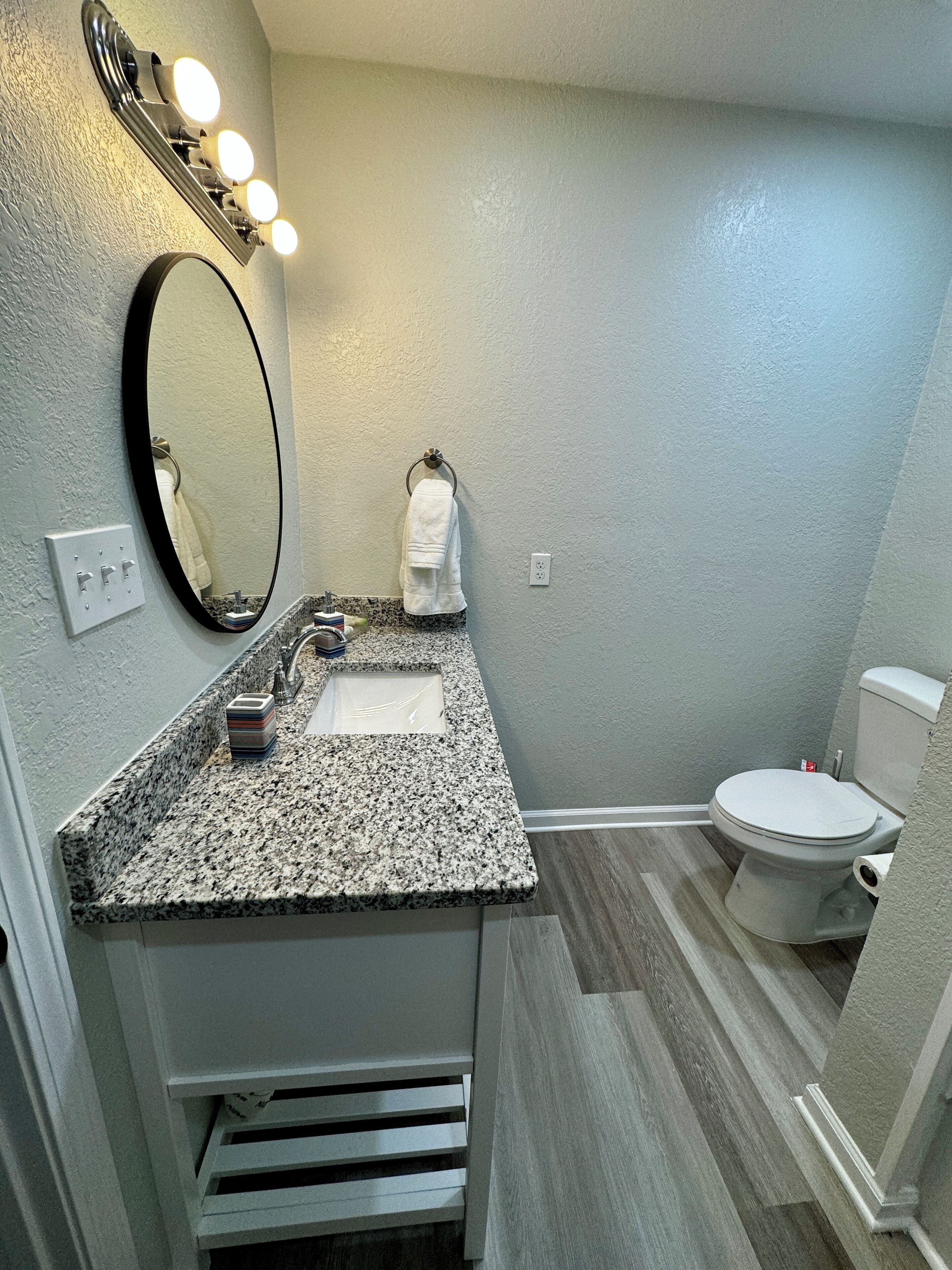 A bathroom with a granite countertop sink, a large mirror with lights, a towel rack, and a toilet.