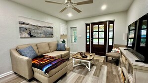 A living room with a beige sofa, a coffee table, a ceiling fan, a painting on the wall, and a sliding glass door leading outside. - Charming 2-bedroom cottage with full kitchen and WiFi in Pensacola. Sleeps 4. (Pensacola)