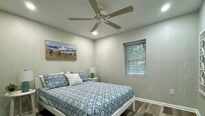 A bedroom with a bed, a ceiling fan, a nightstand with a lamp, a wall-mounted painting, and a window with blinds. - Charming 2-bedroom cottage with full kitchen and WiFi in Pensacola. Sleeps 4. (Pensacola)