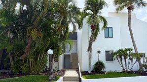 A modern two-story building with a white exterior, a concrete staircase leading to the entrance, and lush tropical landscaping.