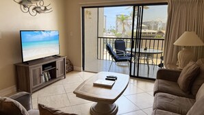 Ein Wohnzimmer mit Fernseher, Couchtisch, Sofa und einem Balkon mit Blick auf den Strand und Palmen.