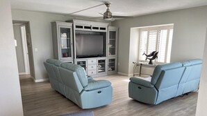A living room with a grey entertainment center, two grey sofas, a ceiling fan, and a dining area with a table and chairs.