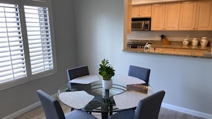 A dining area with a glass table and four chairs, a potted plant, and a kitchen with wooden cabinets and a microwave.