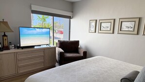 A hotel room with a bed, a TV displaying a beach scene, a chair, a desk with a lamp, and framed pictures on the wall. - Effortless Escape – Walk-Out 2BR at Tamarind (Englewood)