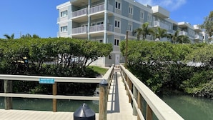 Une jetée en bois menant à un bâtiment doté de nombreux balcons, entouré de verdure et d’eau.
