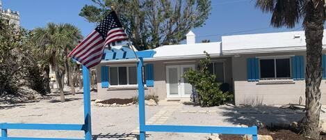 A two-story building with a white facade and blue shutters, surrounded by palm trees and a clear blue sky.