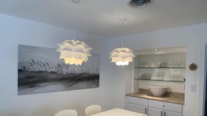 A modern dining area with a white table, chairs, and pendant lights. A large painting of a landscape is on the wall.
