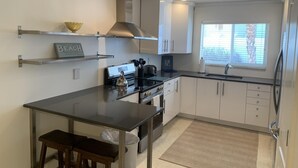 A modern kitchen with white cabinets, a black countertop, and stainless steel appliances. There is a window with blinds, a stainless steel sink, and a small island with stools.