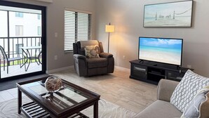 A living room with a sofa, a coffee table with decorative items, a television, and a painting on the wall. - Modern Comfort – 2BR Bayside Condo at Tamarind (Englewood)