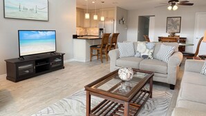 A modern living room with a flat-screen TV, a glass coffee table, and a beige sofa. There is a kitchen area with wooden cabinets and a dining table with chairs. - Modern Comfort – 2BR Bayside Condo at Tamarind (Englewood)