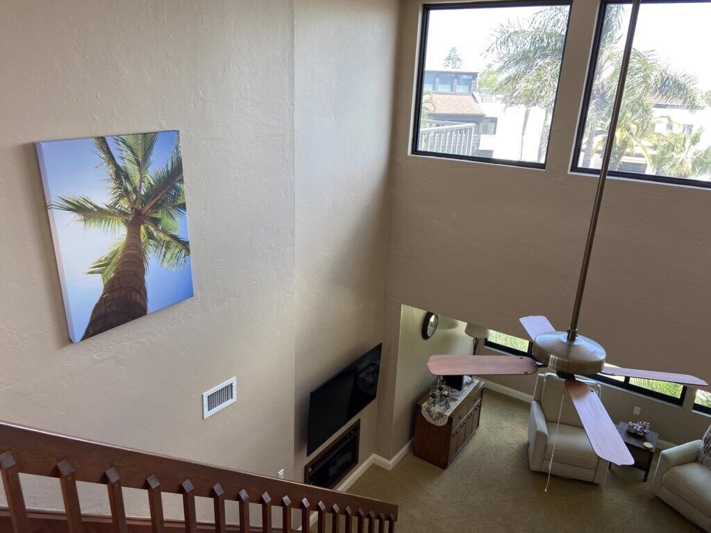 A room with a large window, a ceiling fan, a flat-screen TV, and a painting of a palm tree.