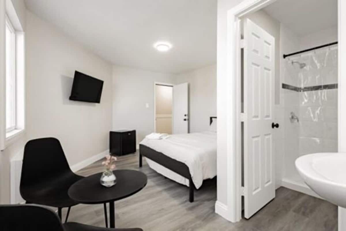 A modern bedroom with a bed, a small table with a vase, a TV on the wall, and a bathroom with a shower.