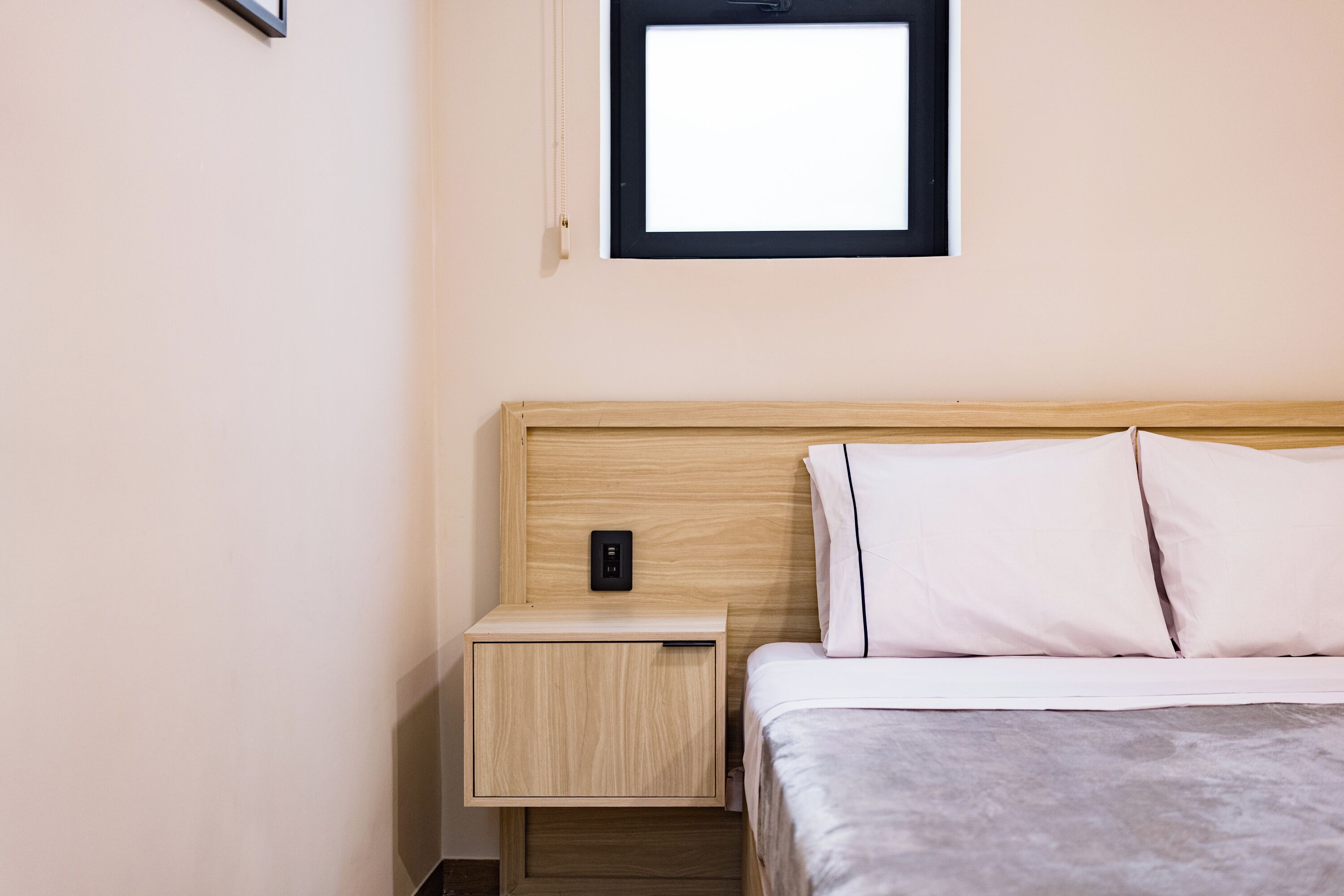 Un dormitorio con cabecera de madera, una mesita de noche con toma de corriente y una ventana sobre la cama.