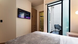 Una habitación de hotel moderna con una cama, un televisor montado en la pared, un armario de madera y un puerta corredera de vidrio que da a un balcón con una silla.