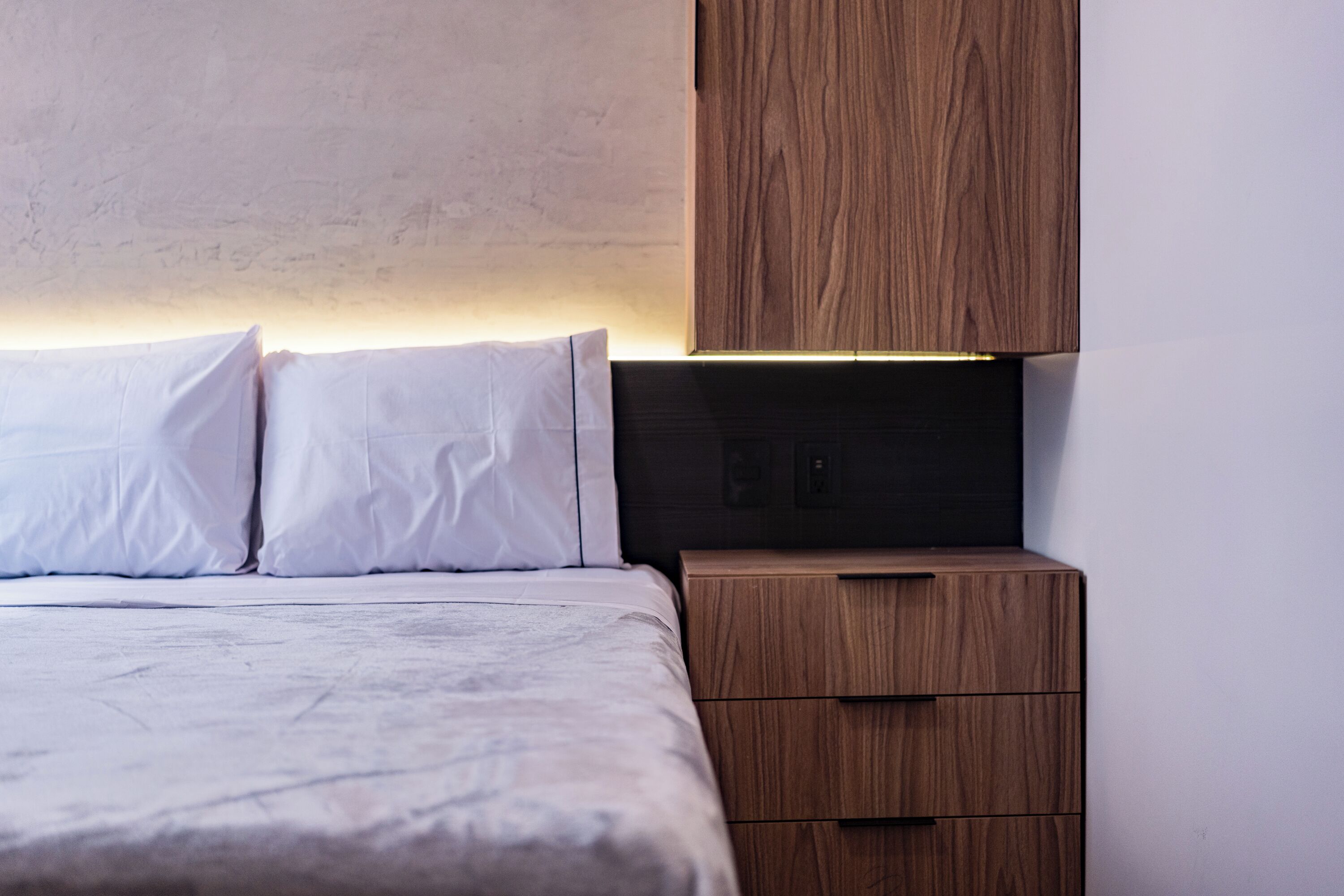 A bed with white linens, a wooden nightstand with drawers, and a wall-mounted cabinet.