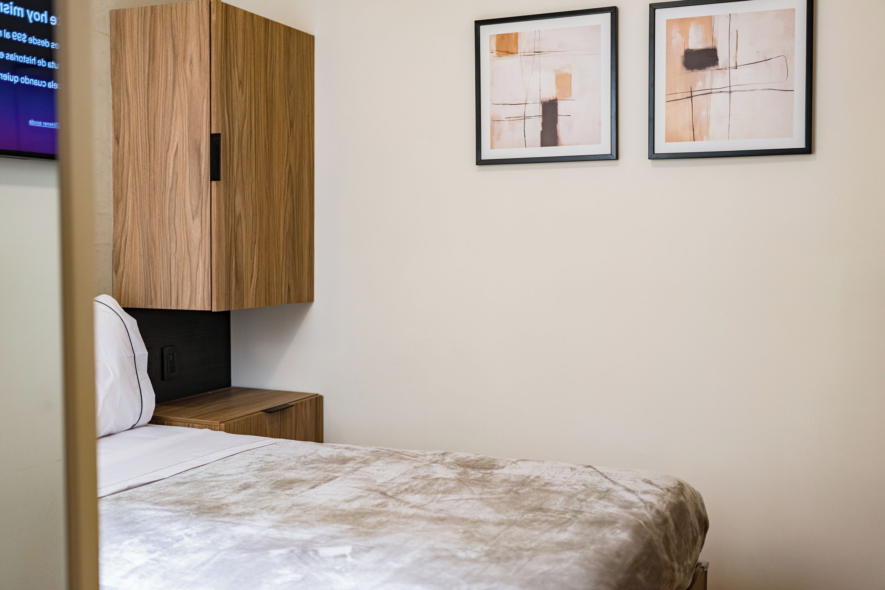 A bedroom with a bed, a wooden wardrobe, a small bedside table, and two abstract paintings on the wall.