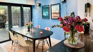 A modern dining area with a dark wooden table, white chairs, and a glass vase with flowers. - Townhouse by Notting Hill - Sleeps5/petsok/tv/wifi (London)