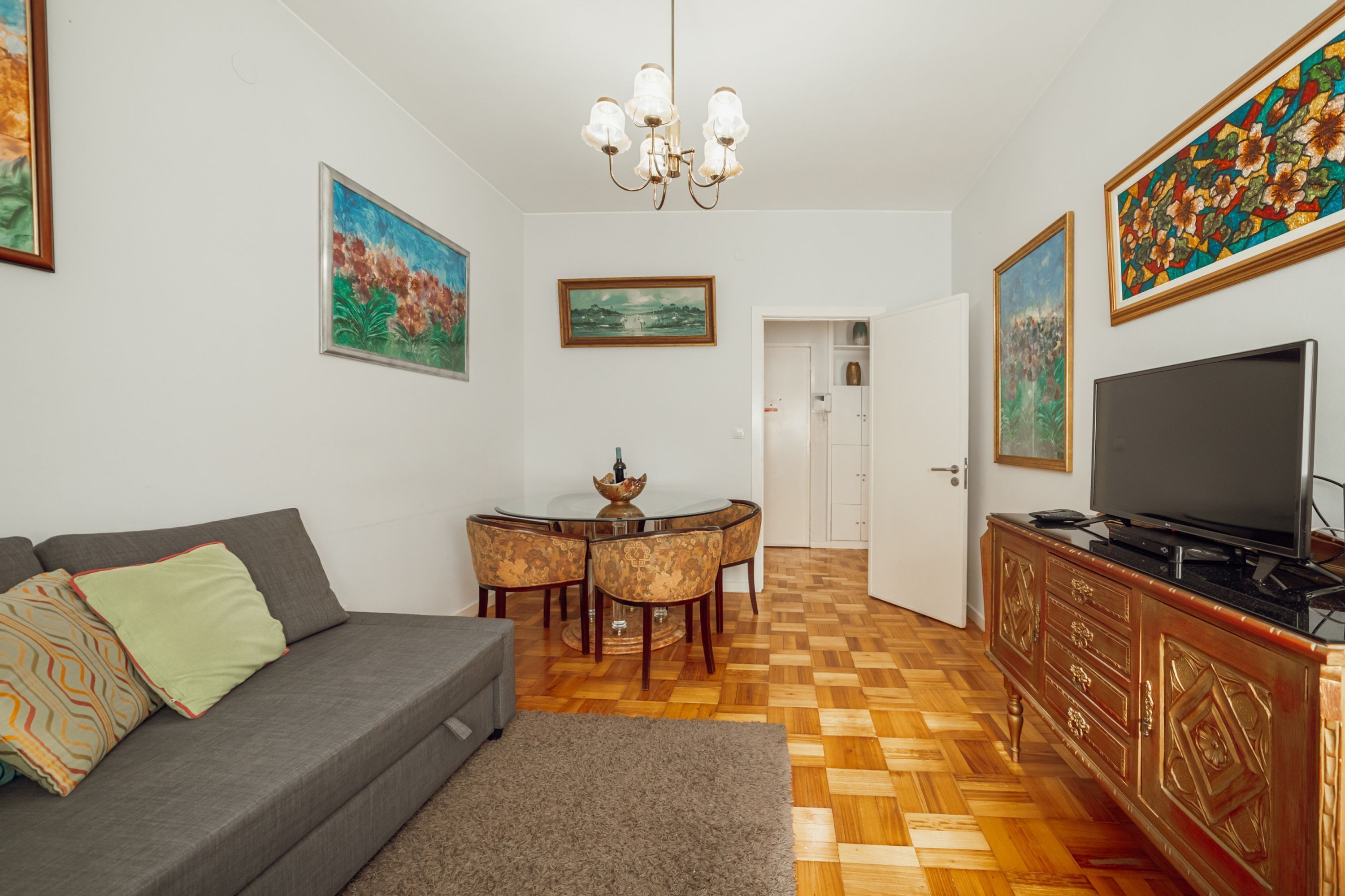 A living room with a sofa, a dining table with chairs, a TV on a cabinet, and artwork on the walls.
