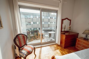 A bedroom with a wooden floor, a chair, a dresser, a mirror, and a balcony with a view of buildings. - Downtown Aquarelle With AC by Vacationy (Porto)