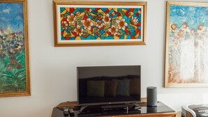 Ein Zimmer mit einem Flachbildfernseher auf einer Holzeinrichtung, darüber ein gerahmtes Blumenbild und ein großes Fenster mit Blick auf Bäume.