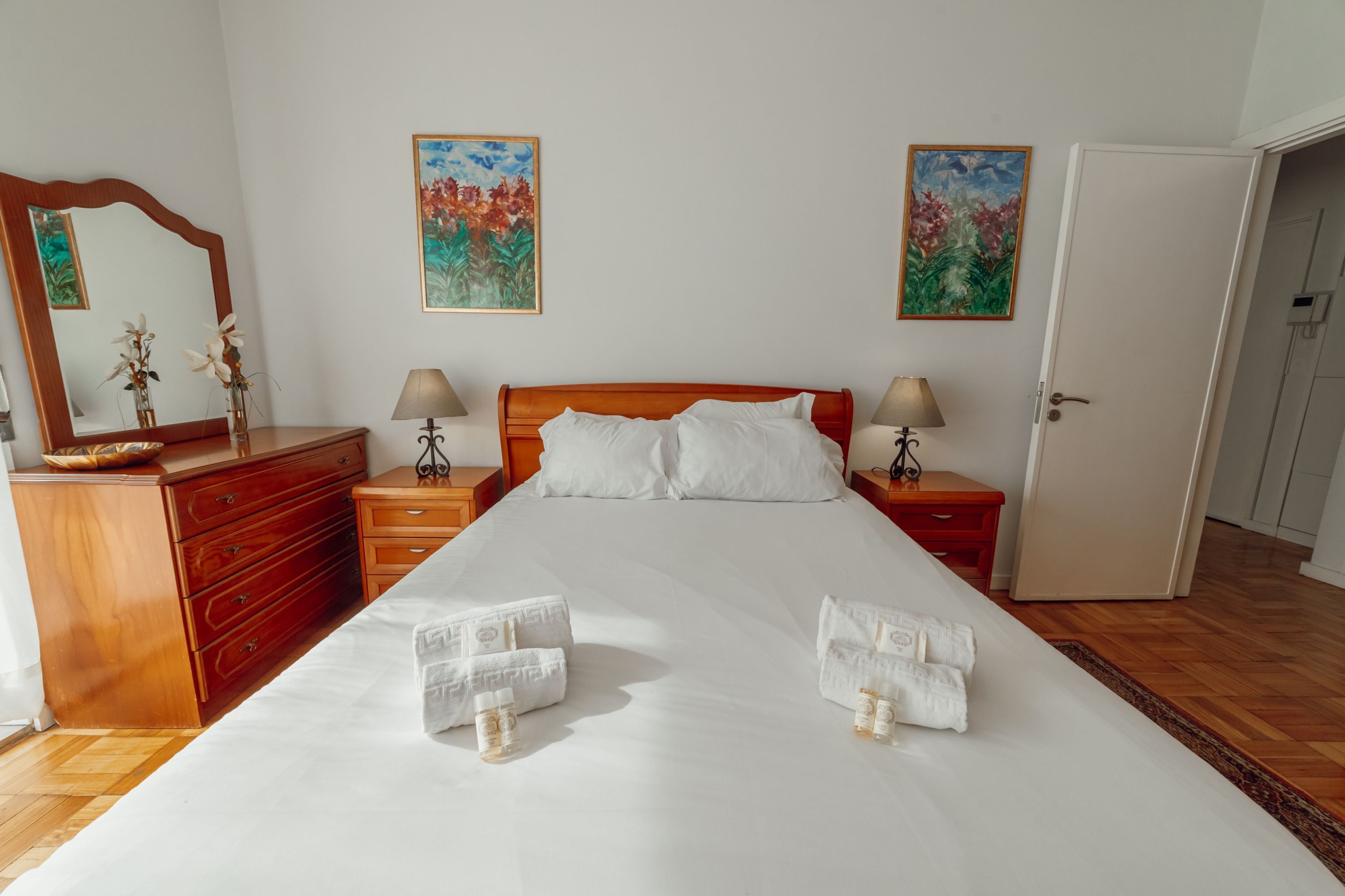 Un lit double bien fait, avec du linge de lit blanc, deux tables de chevet avec des lampes, une commode en bois et deux tableaux aux motifs floraux accrochés au mur.