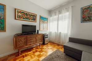 Ett vardagsrum med trägolv, en grå soffa, en tv placerad på ett träskåp och tre inramade tavlor med blommotiv på väggen.