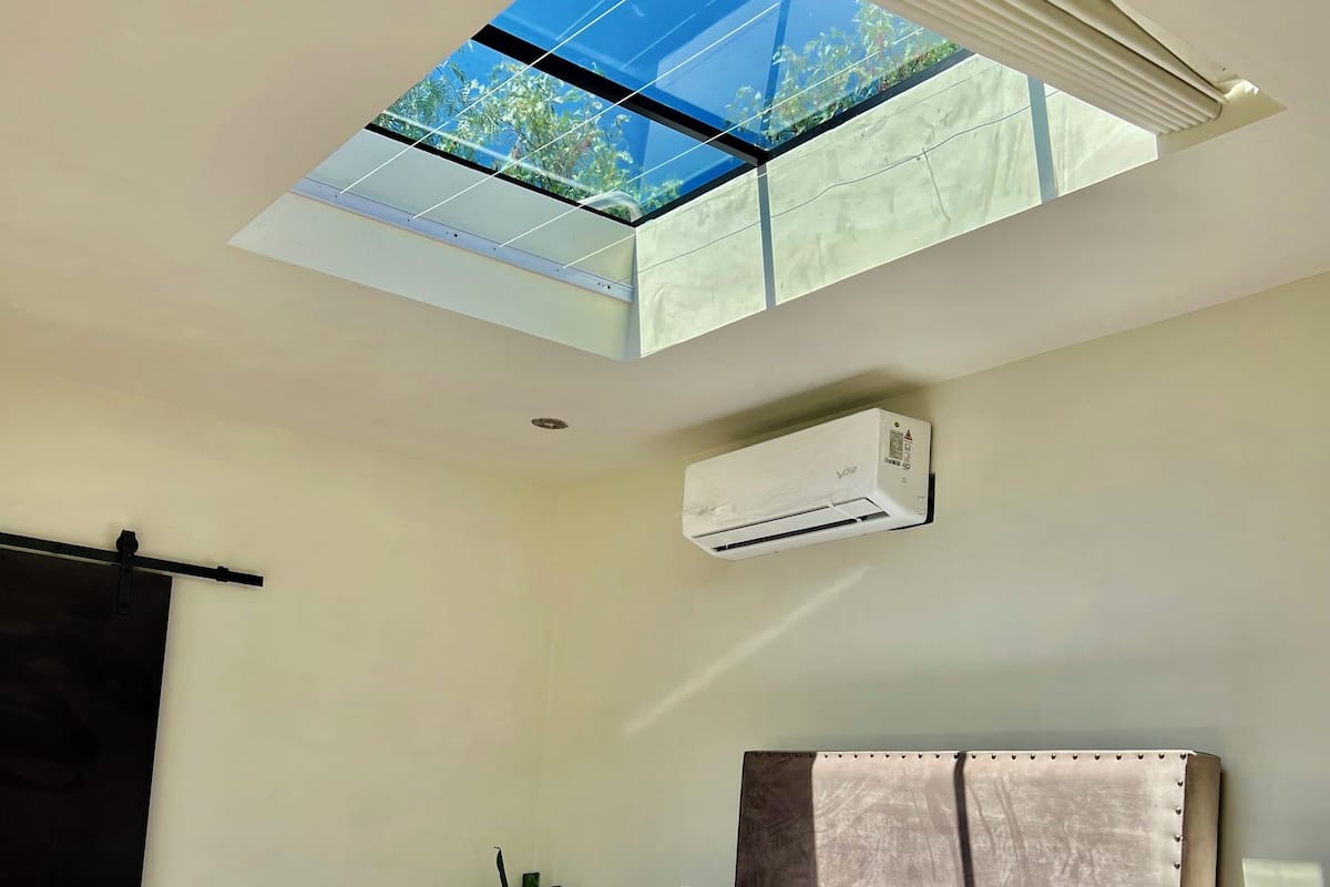 A bedroom with a skylight, a bed with pillows, a bedside plant, and an air conditioning unit.