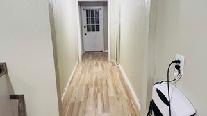 A hallway with wooden flooring, a white door at the end, and a wall-mounted electrical outlet.
