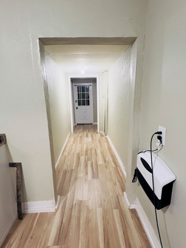A hallway with wooden flooring, a white door at the end, and a wall-mounted electrical outlet.