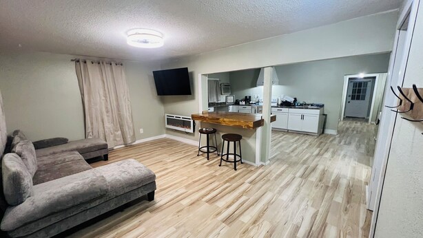 A modern living room with a sofa, a wooden dining table with stools, a kitchen area with cabinets and a TV, and a hallway leading to a door.