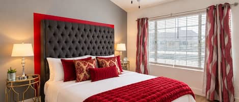 Une chambre d’hôtel avec un grand lit, deux lampes de chevet, un ventilateur de plafond, une fenêtre avec des rideaux et une petite table sur laquelle se trouvent une lampe et une plante.