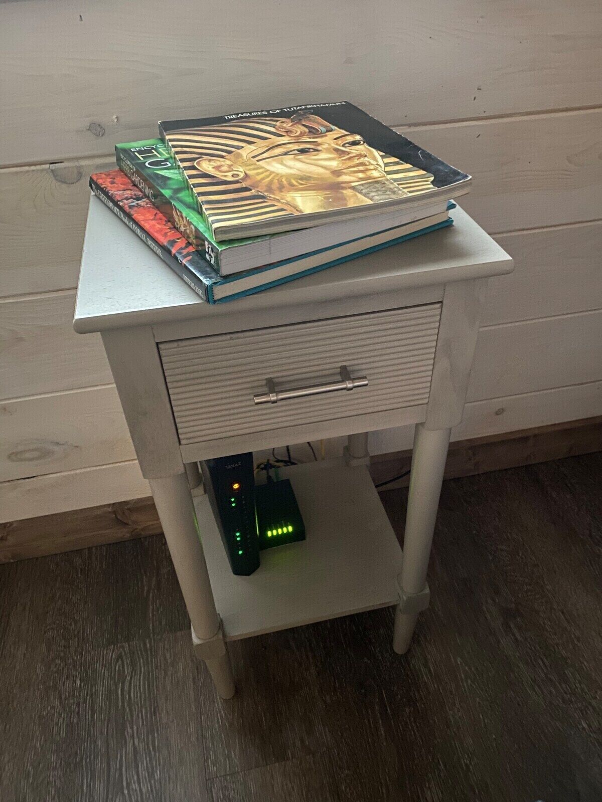 A white nightstand with a drawer and a shelf holding books and a remote control.