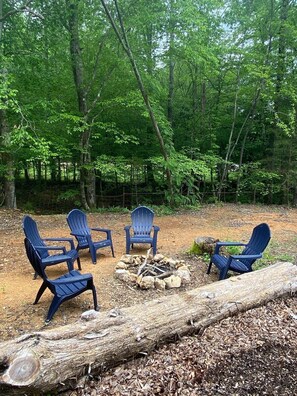 Un terrain de camping boisé, avec quatre chaises Adirondack disposées autour d’un foyer central.