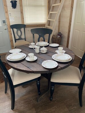 Ein runder Holztisch mit tellerförmigen und becherartigen Geschirren mit Blumenmuster, umgeben von dunklen Holzstühlen mit gepolsterten Sitzen.