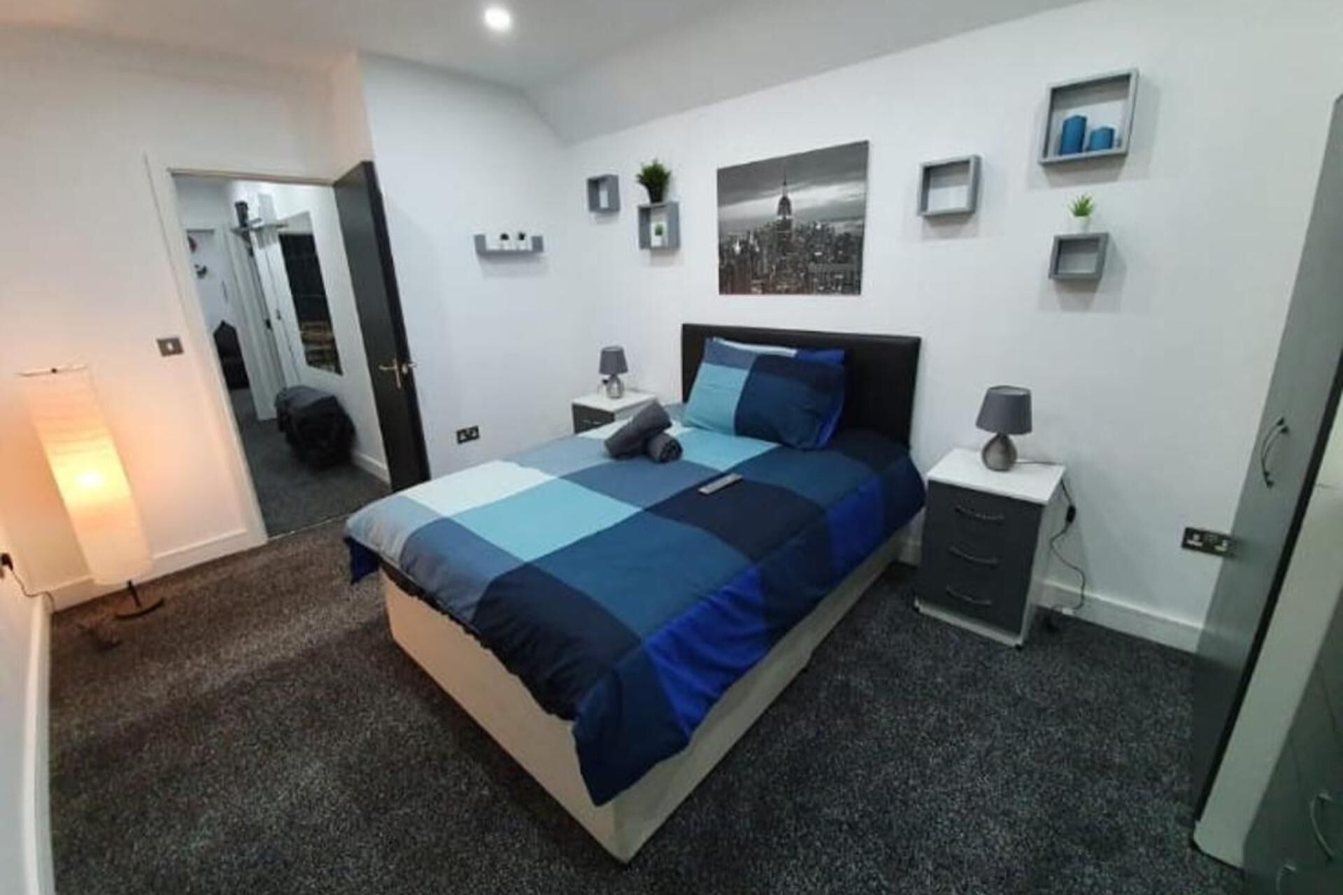 Un dormitorio moderno con una cama grande, mesitas de noche, un espejo y una fotografía de un paisaje urbano en la pared.
