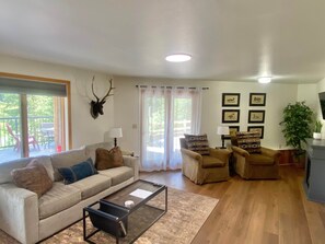 A living room with a sofa, armchairs, a coffee table, a mounted deer head, and a sliding glass door leading to an outdoor area. - Whitefish Riverfront Retreat — 2bd, 1ba on 5 acres (Flathead County)