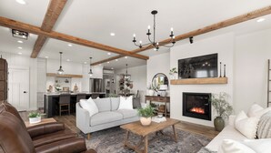 A spacious living room with a fireplace, a sofa, a coffee table, and a dining area in the background. - Gather in Comfort: Large Property with 5 Rentals (Flathead County)