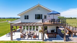 A large white house with a covered patio, outdoor furniture, and a wooden deck. - Gather in Comfort: Large Property with 5 Rentals (Flathead County)