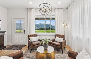 A living room with two leather sofas, a round coffee table, and a view of mountains through large windows. - 2br Cottage w/ Stunning Mountain Views! "Sweetgrass" (Flathead County)
